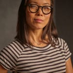 Asian woman with black reading glasses wearing a striped black and taupe shirt posing for a picture.