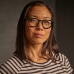 Asian woman with black reading glasses wearing a striped black and taupe shirt posing for a picture.