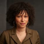 A curly haired black woman dressed in a brown casual jacket.