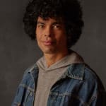 A man with curly black hair wearing a denim jacket looking at the camera.
